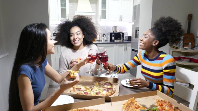 4K视频三位年轻女性坐在厨房里，吃披萨、喝葡萄酒、开玩笑、大笑