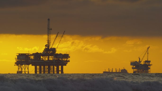 慢镜头拍摄的海浪冲击南加州亨廷顿海滩的海岸，几幅海上石油钻井平台和一艘石油（石油）油轮的剪影在日落时