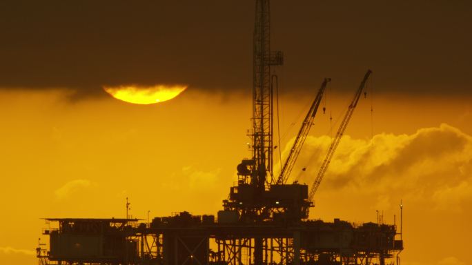 太阳从风暴云下露出，在戏剧性的暴风雨天空下日落时，前景中的海上石油钻井平台的剪影