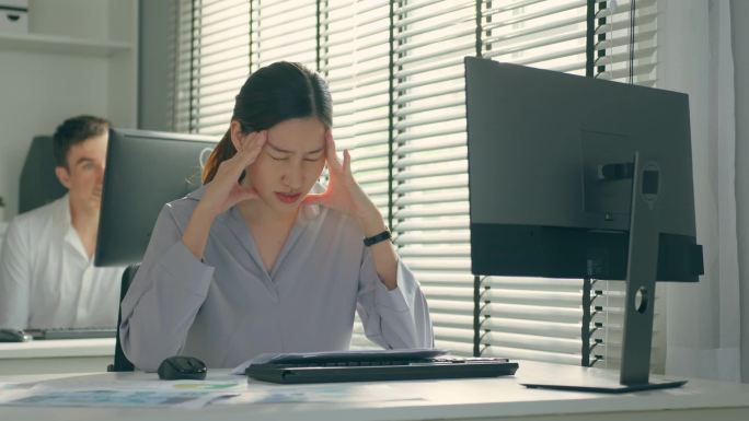 疲惫的亚洲患病商业女工在办公室的桌子上工作。忙碌的员工由于工作迟到和工作过度而感到疲倦和头痛，工作文