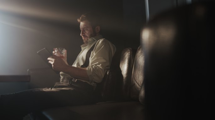 一个穿着衬衫和背带裤的年轻商人坐在黑暗房间的沙发上，看着平板电脑上的东西，喝着威士忌
