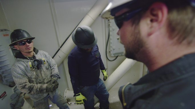 三名男性油田工人在油气钻井平台现场的泵房内交谈并准备打开/关闭阀门以改变泥浆流压力的慢镜头