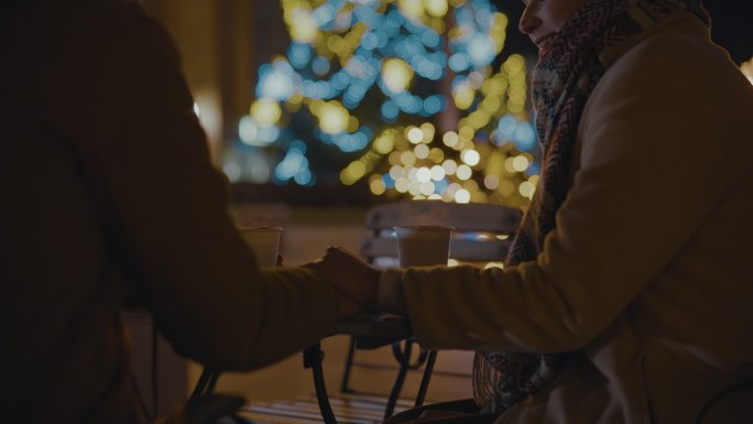 SLO MO一位年轻女子握着男友的手，两人坐在路边咖啡馆的桌子旁，背景是节日的灯光