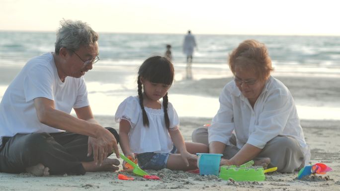 快乐的爷爷，奶奶和孙女在沙滩上的沙盒里为孩子们玩玩具。家庭，生活方式，人，老年人，人寿保险，假期，关