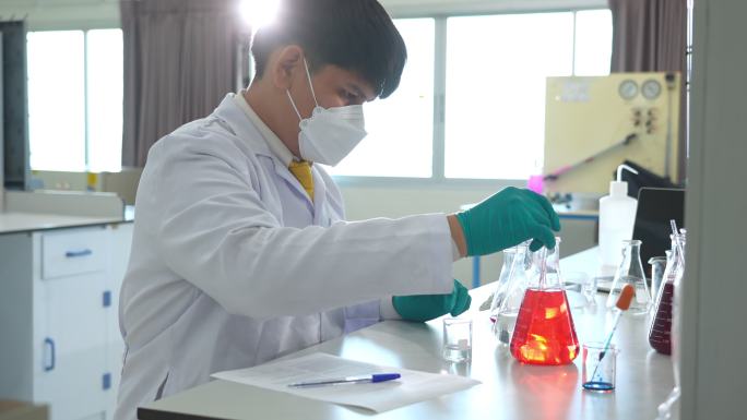 一名亚洲男性医学科学家，穿着实验室工作服和防护设备，仔细检查、测量并使用液体，寻找潜在的新冠肺炎疫苗