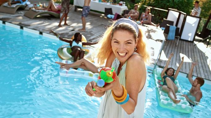 SLO MO金发女子在泳池派对上拿着水枪，对着相机镜头喷水