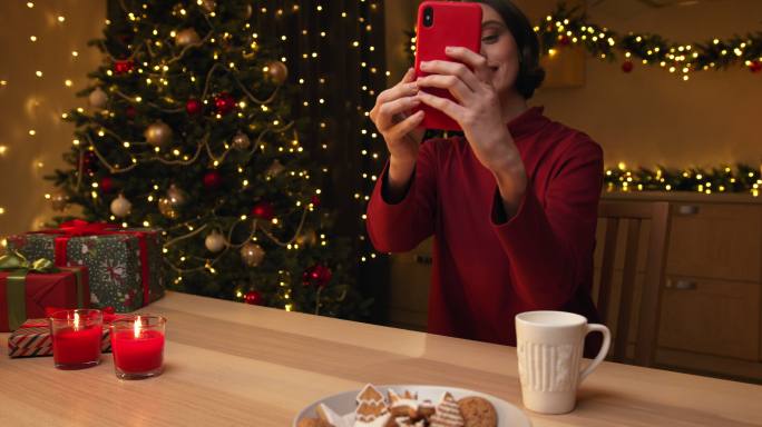 一位身穿红色毛衣的年轻美女面带微笑地坐在桌旁用红色智能手机自拍