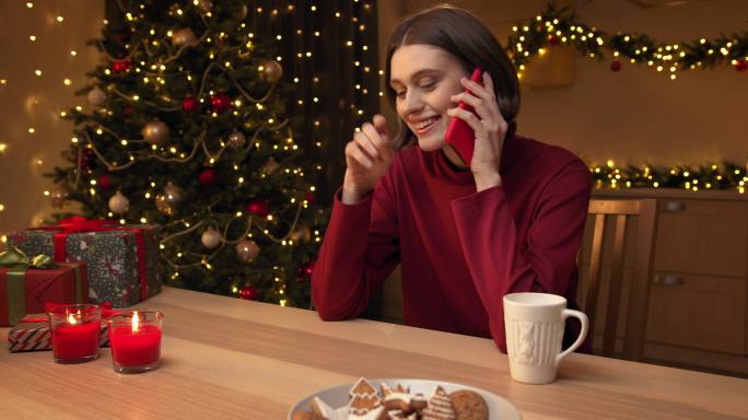 一位穿着红色毛衣的年轻美女正在打电话，然后面带微笑地用智能手机交谈，她坐在桌旁，手里拿着蜡烛