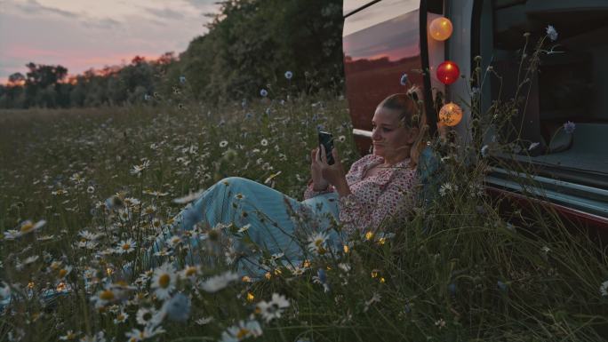 黄昏时分，DS Woman躺在停在草地上的露营车旁，一边使用智能手机