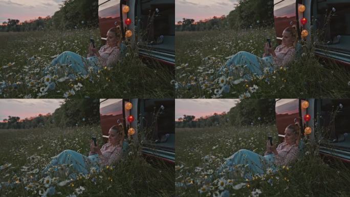 黄昏时分，DS Woman躺在停在草地上的露营车旁，一边使用智能手机
