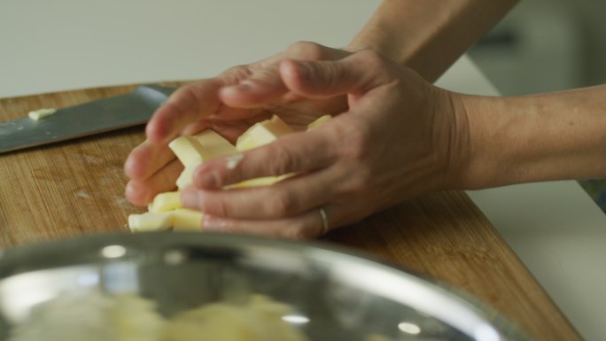 一个年轻女人的手在木制砧板上用一把大菜刀把黄油切成小块，然后拿起黄油撒在金属搅拌碗里的面粉上