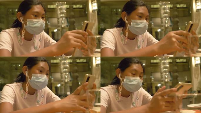 亚洲女孩戴着口罩，在餐厅等待食物时使用智能手机，生活方式理念。