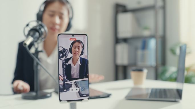 年轻的亚洲女商人在录音室办公室用智能手机录制内容，并在笔记本电脑上播放播客。