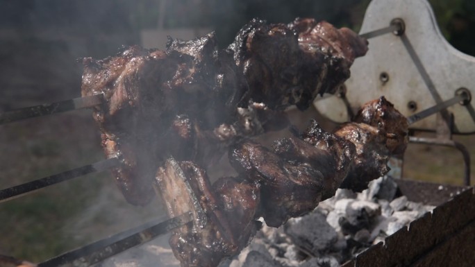 三个缓慢旋转的烤串的斜视图，羊肉被烟雾笼罩，在露天木炭上烹饪时滴落