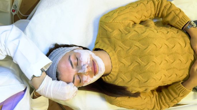 戴着口罩和手套的专业美容师使用专用刷子在女性脸上涂抹嫩肤面膜。库存视频