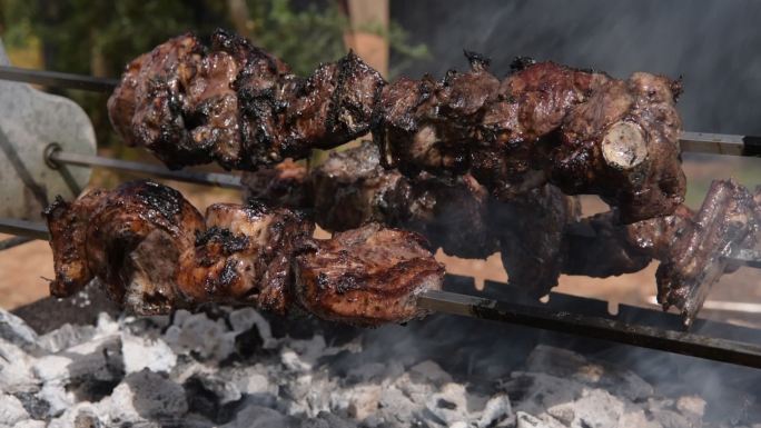 三个缓慢旋转的烤串的斜视图，羊肉在露天木炭上烤时冒着烟，滴水