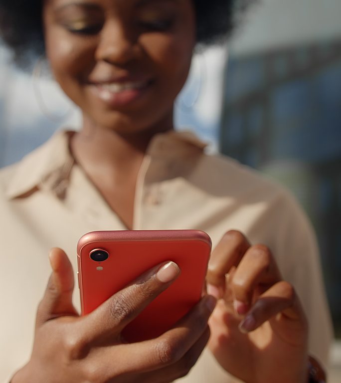 一位身穿米色服装的深色皮肤的漂亮女学生微笑着用红色智能手机发短信，她的脸不在镜头的焦点上，只有手中的