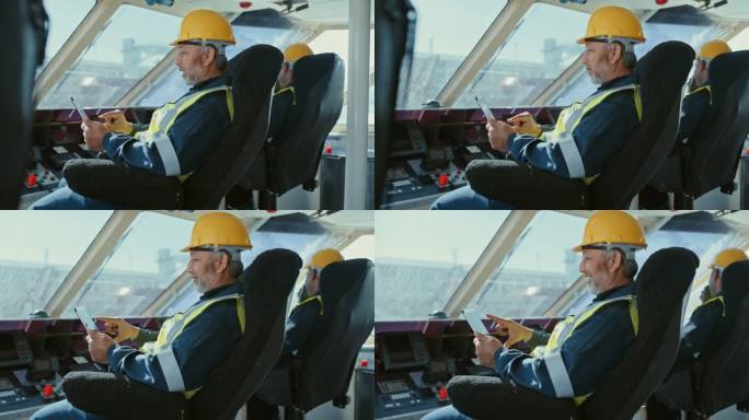 船上两名男子手持数字平板电脑值班。快乐的男性机械师穿着黄色背心，戴着安全头盔，坐在一艘俯瞰大海的商船