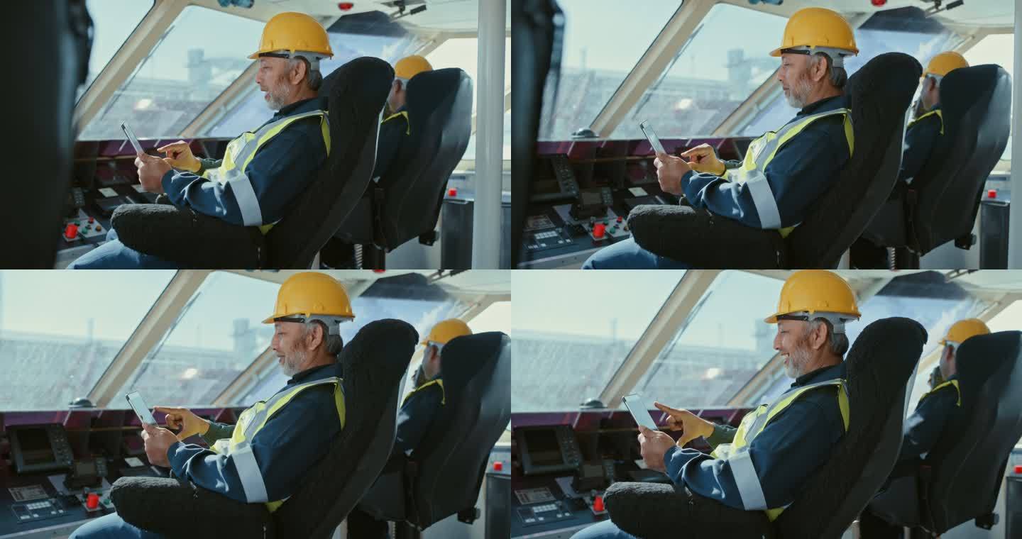船上两名男子手持数字平板电脑值班。快乐的男性机械师穿着黄色背心，戴着安全头盔，坐在一艘俯瞰大海的商船