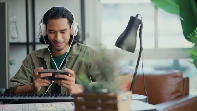 聪明的亚洲男性创意自由职业者休闲布呆在家里坐在工作桌前玩流媒体游戏在线戴耳机微笑无忧无虑的面部表情，