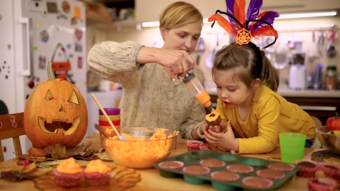好奇的女儿观察着她的母亲，她用绳子上的橙色糖霜装饰万圣节纸杯蛋糕