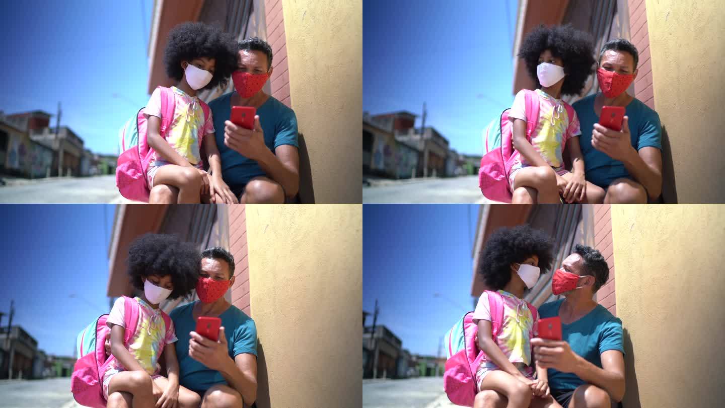 父亲和女儿在家门口等校车并使用手机-带口罩