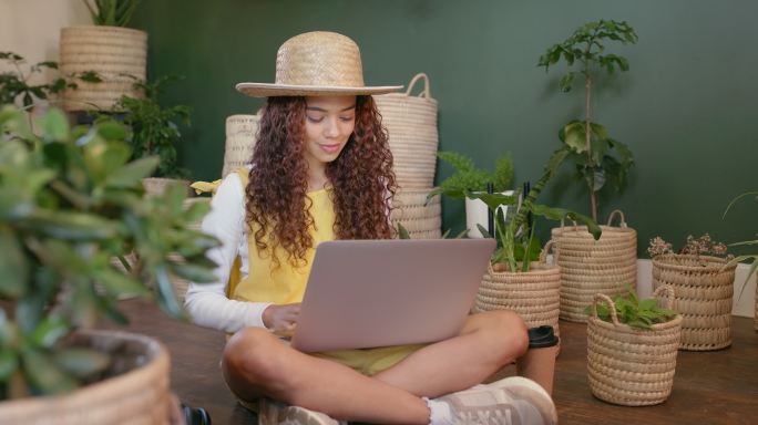 花艺师拿着笔记本电脑，坐在一个充满郁郁葱葱的绿色植物、幼苗和树苗的花园苗圃室里。痴迷于绿色植物的年轻