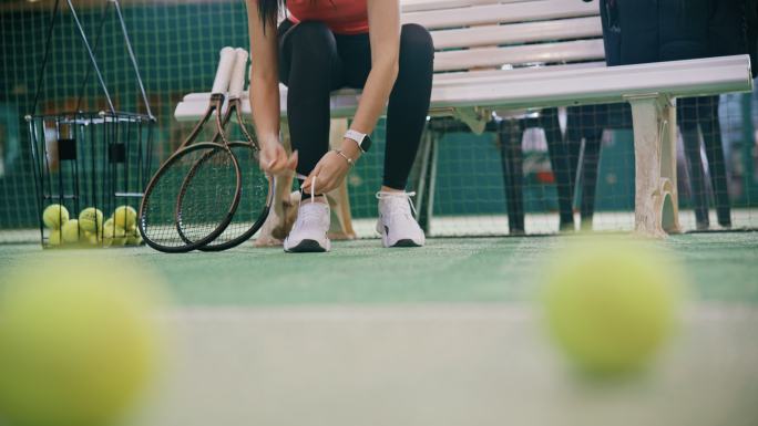 在室内网球场，坐在长椅上系鞋带的年轻女子