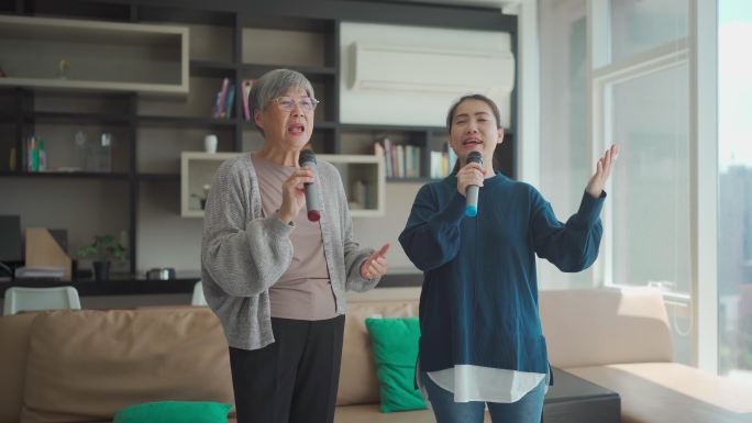 在周末休闲活动中，一名亚洲华裔老年女子在客厅与女儿一起唱卡拉OK跳舞