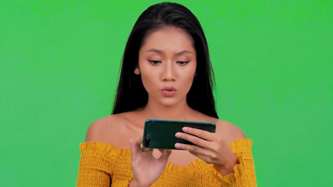 惊讶的年轻黑人女性使用智能手机隔离在绿色背景上。一个快乐的黑人女性用手机庆祝的肖像隔离在绿色的背景上