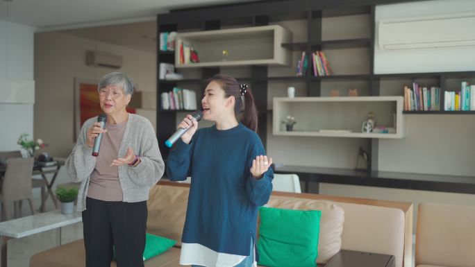 在周末休闲活动中，一名亚洲华裔老年女子在客厅与女儿一起唱卡拉OK跳舞