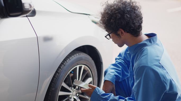 亚洲中年汽车工程师身着蓝色机械连体裤，使用智能手机显示屏检查轮胎气压