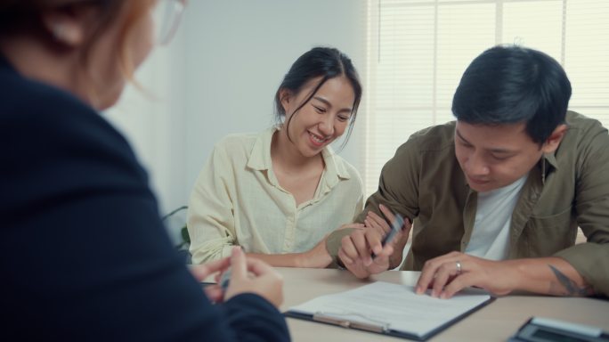 女售货员把房子钥匙交给年轻的亚洲已婚夫妇，他们随意地坐在办公桌前，对在房产办公室签署新的房屋合同感到