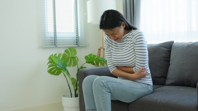 亚洲美丽的生病女孩在客厅里胃痛。迷人的年轻女性在床上醒来后感觉不好，感到疼痛，胃痛或经期，然后在早上