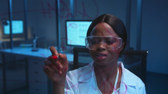 一位年轻的深色皮肤的女化学家穿着实验室外套，在实验室的玻璃板上写下化学公式