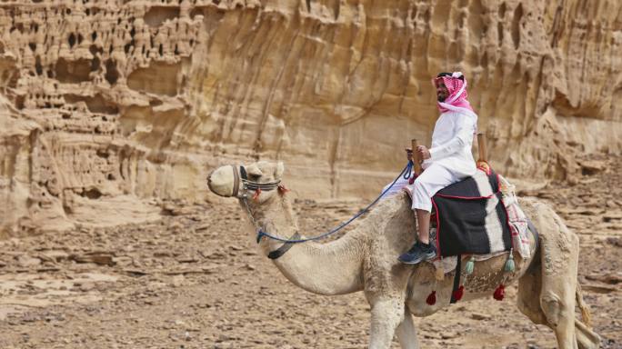 微笑的沙特男子在沙漠地区骑骆驼