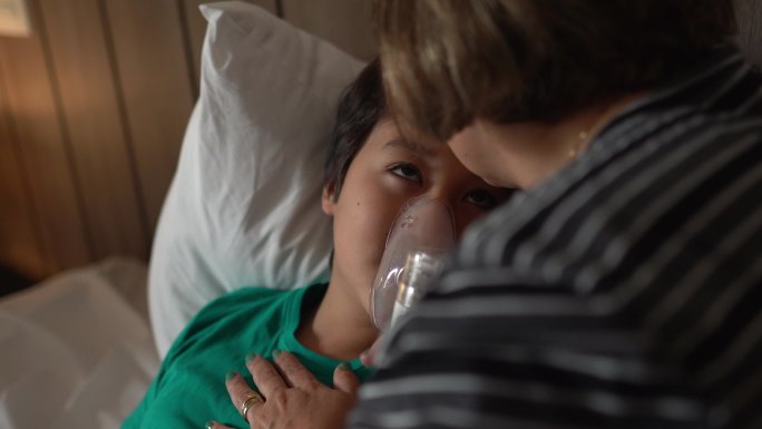 母亲在家中的床上帮助儿子使用喷雾器进行吸入治疗