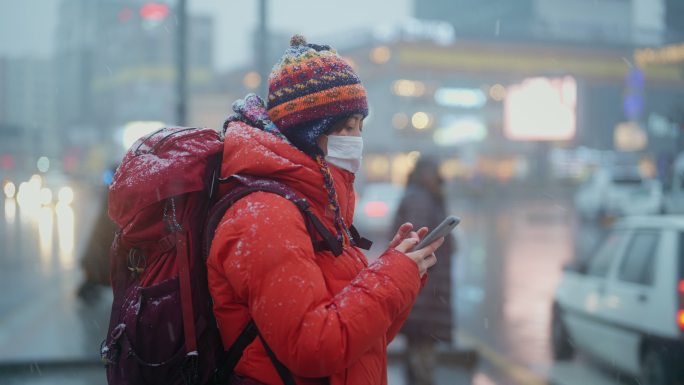 冬天，年轻的女性旅行者使用智能手机在城市的红绿灯前等待