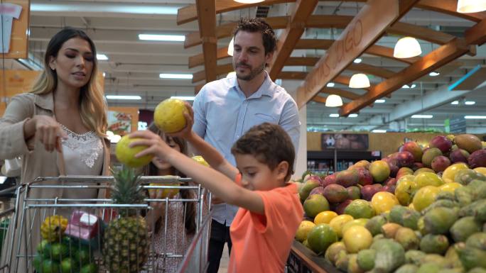 快乐的拉丁美洲家庭在超市购买食品杂货，孩子们在挑选水果，并将其加入购物车