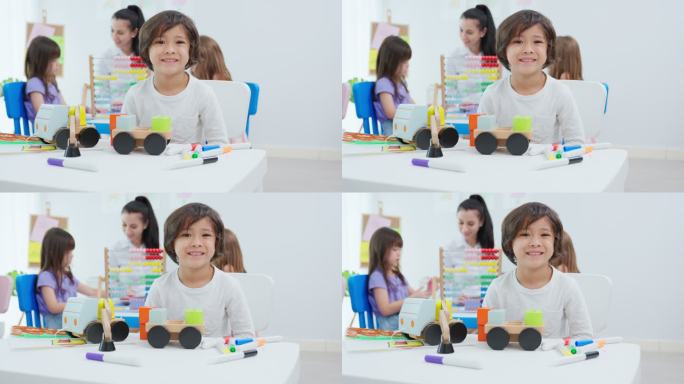 高加索男生在教室里幸福地坐在桌子上的照片。年轻可爱的学龄前儿童看着镜头，美女教师在幼儿园授课