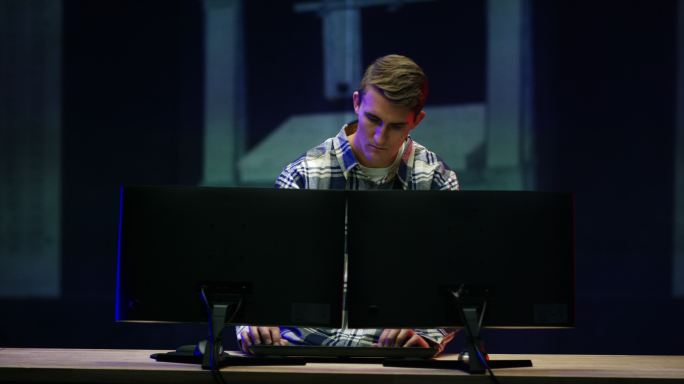 一名20多岁的白人工程师在室内制造设施内用电脑设计3D打印物体，照片中他的设计投影在他身后的墙上