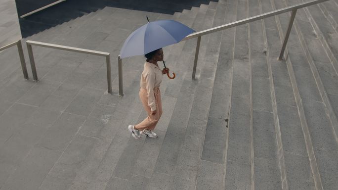 一位年轻迷人的深肤色女商人，身穿米色服装，打着伞，从高处俯瞰，走上楼梯