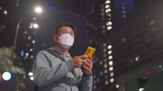 亚洲中国中年男子戴着口罩，在晚上用手机应用程序在线预订后，在城市里举起双臂叫众包出租车