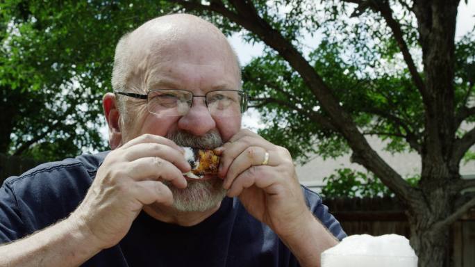 一名50多岁的白人男子拿着啤酒，将煮熟的鸡翅放入牧场敷料中，并在室外吃