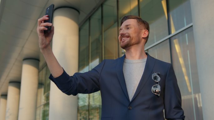 一位身穿蓝色西装、面带微笑的英俊红发青年在现代建筑立面上用手机自拍