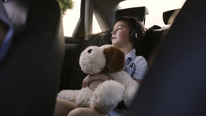 4k视频画面显示，一个小男孩坐在汽车后座上，戴着耳机，紧紧抓住他的毛绒玩具