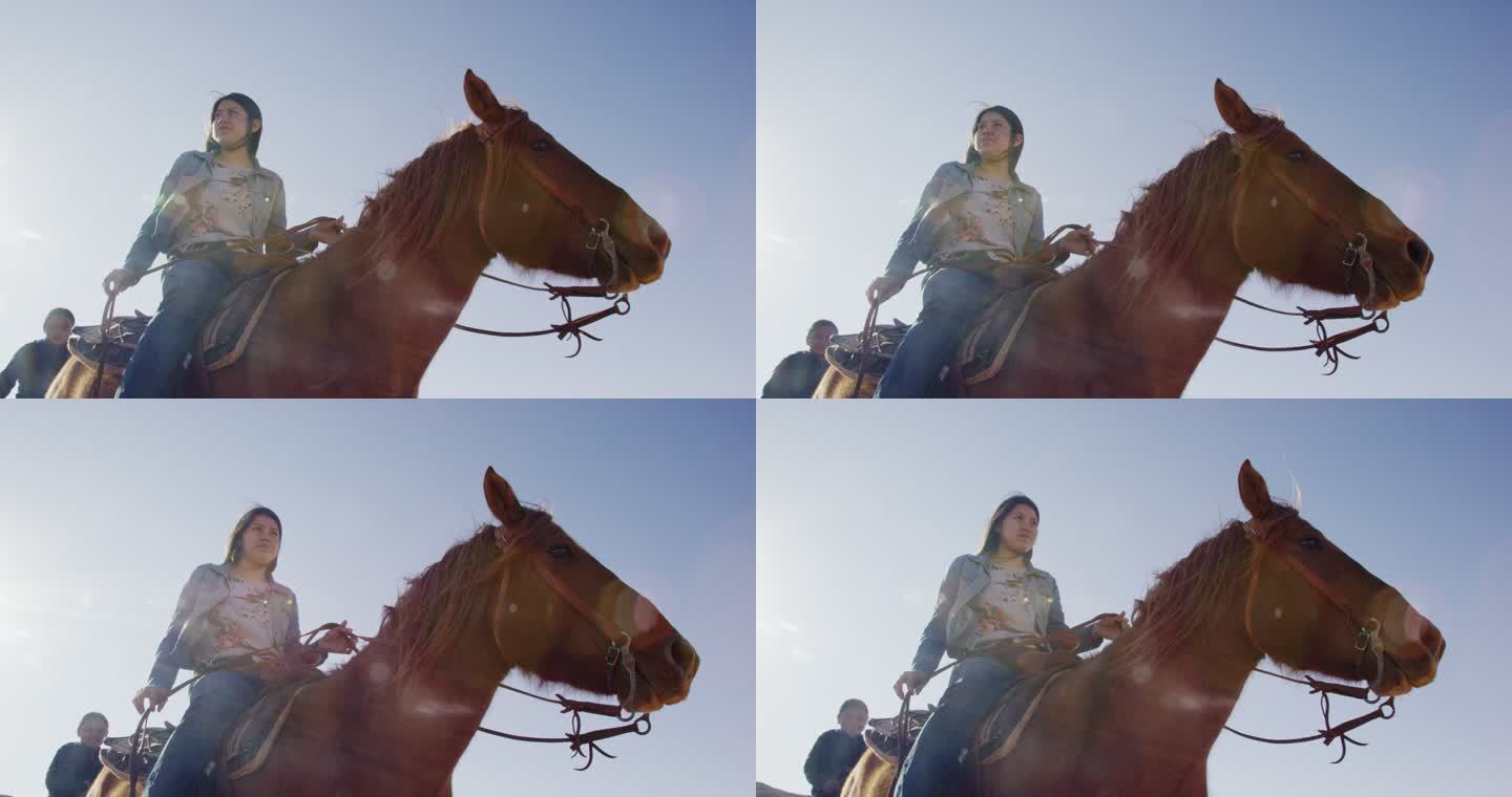 在晴朗明亮的日子里，一位年轻的美国原住民（纳瓦霍族）妇女骑着她的马，身后有一个小男孩