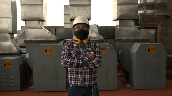 一位拉丁美洲工程师在医院的屋顶上，对着摄像头的风管，戴着防护面罩、头盔和护耳器