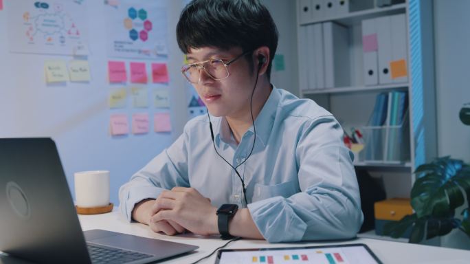 年轻的亚洲商人戴着耳机，使用笔记本电脑和平板电脑观看电脑在线会议演示营销报告数据。