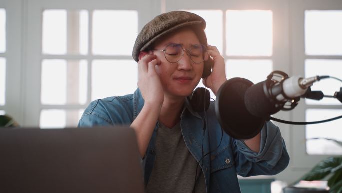 年轻酷炫的亚洲男子手持耳机用电容话筒唱歌，同时在家庭录音室录制新的音乐歌曲